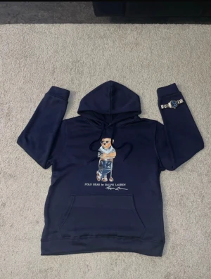 Mörkblå Polo Bear hoodie från Ralph Lauren - Säljer en mörkblå hoodie från Ralph Lauren med Polo Bear-tryck på bröstet. Tröjan har huva med snörning, stor magficka och långa ärmar. Materialet är mjukt och känns som bomull eller polyester. Perfekt för dig som gillar streetwear och snygga prints.