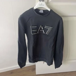 Svart sweatshirt från EA7 Emporio Armani S - Säljer en svart sweatshirt från EA7 Emporio Armani med stort logotryck framtill. Tröjan har rund halsringning och långa ärmar. Perfekt för en stilren och avslappnad look. Materialet känns mjukt och bekvämt mot huden.