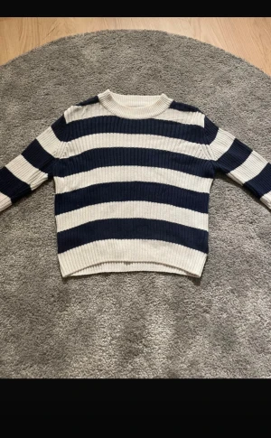 Randig stickad tröja i blått och vitt - Säljer en ribbstickad tröja med breda marinblå och vita ränder. Tröjan har rund hals och långa ärmar, perfekt för dig som gillar klassisk och chill stil. Materialet är mjukt och stickat, vilket gör den skön att bära under kyliga dagar.
