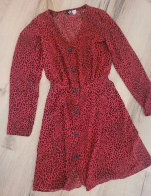 Röd leopardmönstrad klänning från H&M Divided - Röd kortklänning  med svart leopardmönster, v-ringning och knappar framtill. Långärmad och figurnära i midjan för en snygg siluett. Perfekt för dig som vill sticka ut med ett djurmönstrat statement-plagg.