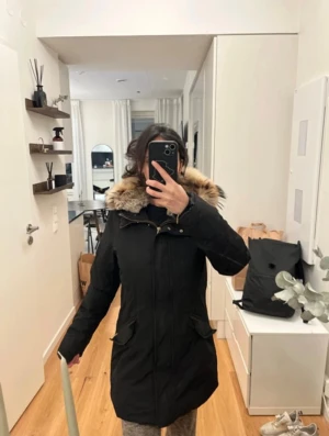 Svart parkajacka med päls från Woolrich - Parkajacka från Woolrich i svart med fluffig beige-brun päls på huvan. Jackan har dragkedja och knappar framtill, två stora fickor med lock och en längre, normal passform. Perfekt för kalla dagar med sin varma och stilrena look.