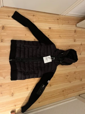 Svart Moncler pufferjacka med huva - Säljer en svart pufferjacka från Moncler med stickade ärmar och huva. Jackan har dragkedja framtill, två fickor med knappstängning och en extra ficka med dragkedja på ärmen. Moncler-logga på vänster ärm och cool quiltad design på kroppen.