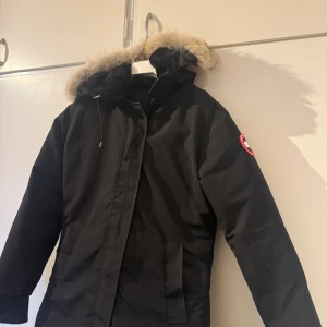 Svart Canada Goose parkajacka M - Svart parkajacka från Canada Goose med ikonisk patch på ärmen och fluffig päls runt huvan. Jackan har två stora fickor framtill, dragkedja och knappar. Perfekt för kalla vinterdagar och har en snygg, klassisk look.