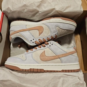 Nike Dunk Low i beige och ljusbrun mocka - Nike Dunk Low sneakers i en snygg mix av beige och ljusbrun mocka med vita detaljer. Skorna har klassisk låg siluett, perforerad tåbox och bruna snören. Swoosh-loggan och yttersulan är i matchande ljusbrunt. Perfekta för dig som gillar stilrena och trendiga sneakers.