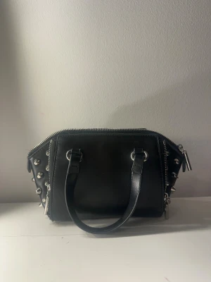 Svart handväska med nitar och dragkedjor - Cool svart handväska i skinn med silverfärgade nitar på sidorna och dubbla dragkedjor upptill. Väskan har två handtag och en rymlig insida med fack. Alla bitar e kvar förutom 1 på sidan (se sista bilden)💓