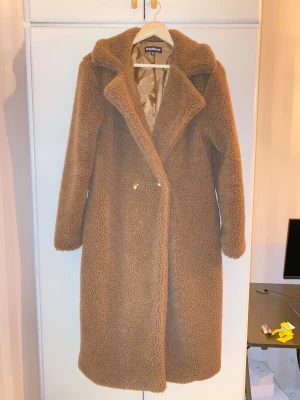 Brun teddyjacka från EillyBazar - Säljer en lång brun teddyjacka från EillyBazar med bred krage och snygga mönstrade knappar. Jackan har ett mjukt och fluffigt material, helfodrad insida i satin och klassisk rak passform. Perfekt för dig som vill ha en cozy och trendig look i höst och vinter. Den är helt ny min mamma köpte den och tyckte att den va för stor för henne så den ba låg i garderoben  ny pris för 669. 