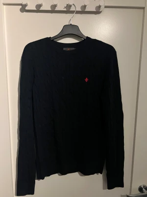 Mörkblå 100% merinoull kabelstickad Morris tröja - Svart kabelstickad tröja från Morris med rund hals och lång ärm. Tröjan har en liten röd logga broderad på bröstet och är gjord i mjuk bomull. Perfekt för dig som gillar klassisk stil med en modern touch. Kvitto och allt tillbehör följer med. 