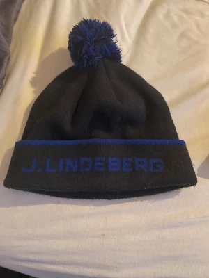 Svart mössa med blå tofs J.Lindeberg - Snygg svart stickad mössa från J.Lindeberg med blått broderat logotyp på uppviket och en fluffig blå tofs på toppen. Mössan har en blå rand längs kanten och är gjord i mjukt material som håller dig varm. Perfekt för kalla dagar.