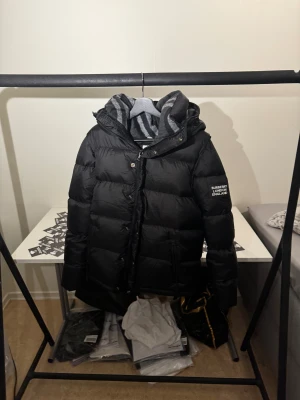 Svart Burberry pufferjacka med avtagbara ärmar - Svart pufferjacka från Burberry med avtagbara ärmar, så du kan rocka den som väst också. Jackan har huva, dragkedja och knappar framtill samt Burberry London England-tryck på ärmen. Fodret är grått med zebramönster. Perfekt för kalla dagar.