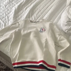 Vit sweatshirt från Moncler med ränder - Vit sweatshirt från Moncler med broderad logga på bröstet. Tröjan har långa ärmar och ribbade muddar med röda, blå och vita ränder. Rund hals och mjukt material som känns skönt mot huden. Perfekt för dig som gillar stilrena och sportiga vibes.