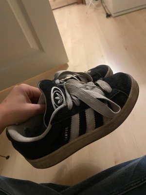 Adidas Campus sneakers svart/vit 38⅔ - Man får med ett par nya sko snören. Svarta Adidas Campus sneakers med klassiska vita ränder och logga på plösen. Skorna har rund tå, platt sula och är tillverkade i mocka med vita snören och detaljer. Perfekt för dig som gillar retrostil och streetwear.