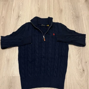 Marinblå stickad tröja Polo Ralph Lauren - Snygg marinblå stickad tröja från Polo Ralph Lauren med half zip och kabelstickat mönster. Tröjan har lång ärm, ribbad krage och muddar samt röd broderad logga på bröstet. Perfekt för dig som gillar klassisk stil med en modern touch.