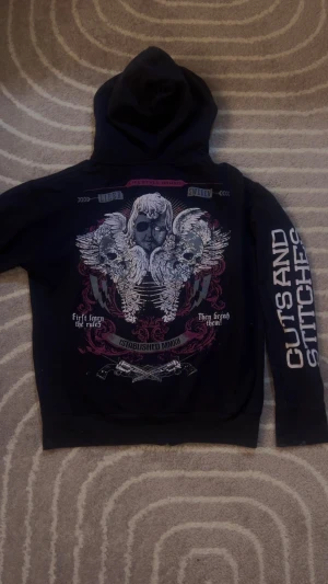 Svart hoodie Cuts and Stitches - Svart hoodie med dragkedja från Cuts and Stitches. Stor tryckt logga på bröstet och ärmen, samt detaljerad rygg med dödskallar, vingar och text. Huva med snörning och långa ärmar. Snygga kontrastdetaljer i vitt och rött. Perfekt för dig som gillar streetwear. Har arm holes