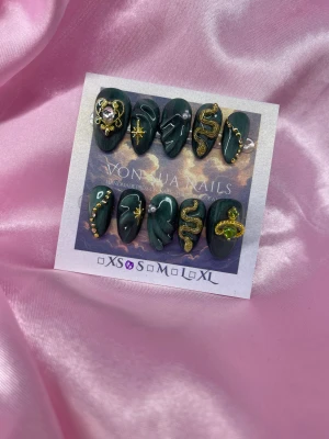 Emeri (S);Short Almond;Curved - Set med handgjorda cateye mörkgröna press-on naglar dekorerade med detaljer i guld, pärlor och gnistrande stenar. Designen har motiv som ormar, stjärnor och hjärtan, samt 3D-effekter och glittriga accenter. Naglarna har en elegant, kort mandel form och passar dig som gillar unik och edgy stil.S: 15 mm, 11,5 mm, 12 mm, 11 mm, 9 mm 
