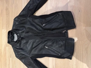 Svart skinnjacka Calvin Klein Jeans - Snygg svart skinnjacka från Calvin Klein Jeans med perforerat mönster och coola detaljer som snedställda dragkedjor och ståkrage med tryckknapp. Jackan har lång ärm och en slim passform som ger en modern look. Perfekt för dig som gillar streetstyle. Aldrig använd, säljer på grund av detta.