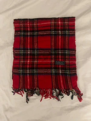 Rutig röd halsduk från Burberrys - Klassisk rutig halsduk från Burberrys i rött, svart, vitt och gult. Tillverkad i 100% lammull och har fransar i ändarna. Perfekt accessoar för att lyfta din vinterstil med en ikonisk brittisk touch.