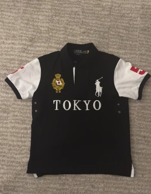 Svart och vit polotopp från Ralph Lauren - Säljer en snygg polotopp från Ralph Lauren med svart kropp och vita korta ärmar. Stora broderade detaljer med texten TOKYO, gul emblem och vit polospelare på bröstet. Röd siffra på ärmen och klassisk krage. Perfekt för dig som gillar sportig och stilren look.