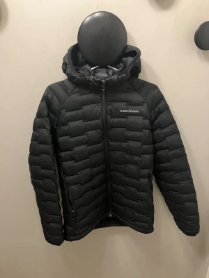 Svart lättviktsdunjacka Peak Performance - Svart dunjacka från Peak Performance med huva och dragkedja framtill. Jackan har quiltad design och logga på bröstet. Perfekt för kalla dagar och har en stilren look som funkar till det mesta.