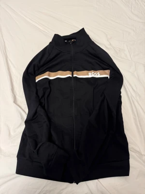 Svart zip hoodie från BOSS - Svart hoodie från BOSS med hel dragkedja och ståkrage. Snyggt brunt och vitt band över bröstet med BOSS-logga. Tillverkad i mjuk bomullsmix, perfekt för chill eller streetwear. Långärmad och med ribbade muddar för extra komfort.