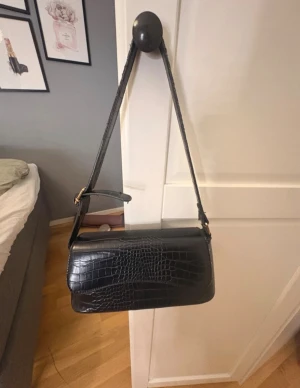 Svart axelväska i croco-mönstrat skinn - Snygg svart axelväska med croco-mönstrad yta och justerbart axelband. Väskan har ett stilrent, avlångt format och stängs med lock. Invändigt finns ett fack med dragkedja för småsaker. Perfekt för dig som gillar trendiga accessoarer med lyxig känsla. Ganska sliten men går fortfarande att använda.