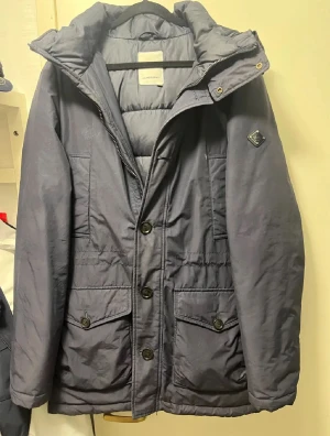 Mörkblå  VinterJacka från J.Lindeberg - Vinterjackan är i mörkblått från J.Lindeberg, med Luva och både dragkedja och knappar framtill. Jackan har två stora fickor med lock och knappar, samt en diskret logga på ärmen. Perfekt för kalla dagar med sin långa modell och fodrade insida.       (SNABB LEVERANS)