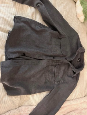 Mörkgrå overshirt från H&M - Snygg mörkgrå overshirt från H&M med två stora bröstfickor och knappar framtill. Skjortan har lång ärm och är tillverkad i ett mjukt, ull-liknande material som ger en chill och stilren vibe. Perfekt att slänga över en t-shirt för en cool look.