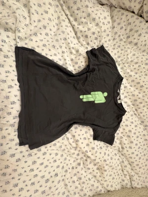 Svart Billie Eilish t-shirt med tryck - Svart t-shirt från Billie Eilish med grönt tryck av en ikon på framsidan och artistens namn i coolt typsnitt på ryggen. Klassisk rund hals och korta ärmar. Mjuk och skön bomull som passar perfekt för dig som gillar streetstyle och musikmerch.