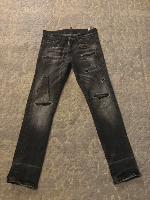 Svarta distressed jeans från Dsquared2 - Säljer ett par svarta jeans från Dsquared2 med coola slitningar och hål på både fram- och baksidan. Jeansen har en straight fit och klassisk femficksdesign. Materialet är jeans med en tvättad look och tydliga slitdetaljer för en edgy vibe. Det finns ett fixat hål som knappast syns