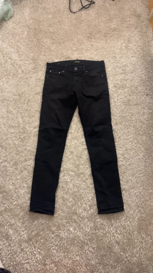 Jacka&Jones Svarta Slim Glenn jeans storlek 30/32 - Svarta jeans från Jack & Jones, modell Slim Glenn. Snygg slim fit med smalare ben och klassisk femficksdesign. Jeansen har knapp och dragkedja i gylfen, och är gjorda i mjukt bomullsmaterial med lite stretch för extra komfort.