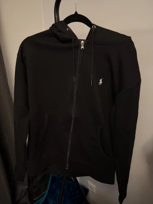 Svart hoodie från Polo Ralph Lauren - Svart hoodie från Polo Ralph Lauren med klassisk logga broderad på bröstet. Tröjan har huva med snörning, dragkedja framtill och två fickor. Materialet är mjuk bomull och modellen är normal passform med långa ärmar. Perfekt för chill dagar.