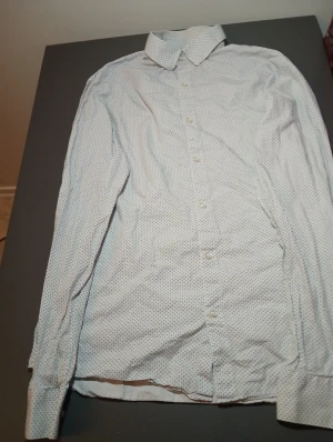 Vit prickig skjorta med knappar - Snygg vit skjorta med små svarta prickar och klassisk krage. Skjortan har lång ärm och knappar hela vägen framtill. Perfekt för dig som gillar stilrena detaljer och ett fräscht uttryck.