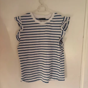 Randig volangtopp från SHEIN  - Säljer en vit och blå randig topp från SHEIN i storlek XS. Toppen har volangdetaljer vid axlarna och är ärmlös, vilket ger en söt och trendig look. Tillverkad i mjuk bomull, perfekt för en fräsch och avslappnad stil.