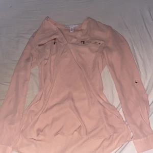 Ljusrosa blus med dragkedjor från Nelly - Säljer en ljusrosa blus från Nelly med v-ringning och dekorativa dragkedjor vid bröstet. Blusen har långa ärmar och är tillverkad i ett lätt och mjukt material som ger en snygg och avslappnad look.