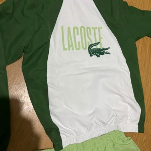 Vita och gröna Lacoste täckbyxor - Säljer ett par vita täckbyxor från Lacoste med ljusgröna och mörkgröna detaljer. Byxorna har resår i midjan och vid bensluten samt snörning. Materialet är syntet, perfekt för sportiga aktiviteter. Klassisk Lacoste-logga på benet.