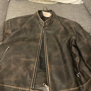 Brunskinnjacka från Nelly, stl S - Cool brun skinnjacka från Nelly i bikerstil med slitna detaljer. Jackan har dragkedjor på fickor och ärmslut, ståkrage och tryckknappar vid midjan. Perfekt för dig som vill ha en edgy look med vintagekänsla.