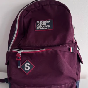 Purple ryggsäck från Superdry - Snygg vinröd ryggsäck från Superdry Japan Outdoor med flera fack, justerbara axelremmar och coola detaljer som randiga dragkedjor och logopatch. Perfekt för skolan eller stan, rymmer laptop och allt du behöver. Materialet är slitstark polyester.