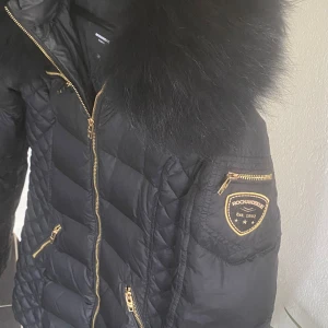 Svart pufferjacka Rockandblue päls - Säljer en svart pufferjacka från Rockandblue med stor fluffig fuskpäls runt huvan och snygga guldiga dragkedjor. Jackan har quiltade partier på sidorna, Rockandblue-märke på ärmen och flera fickor med dragkedja. Perfekt för kalla dagar och riktigt stilren look.