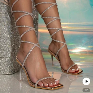 Glittriga sandaletter med snörning - Snygga sandaletter i beige satin med smala remmar dekorerade med strass som snörs upp längs benet. Skorna har fyrkantig tå, öppen tå och hög klack i transparent stil. Perfekta för att sticka ut och ge en lyxig känsla till din outfit. 