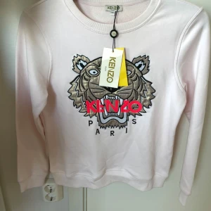 Ljusrosa Kenzo sweatshirt med tiger - Superfin ljusrosa sweatshirt från Kenzo med ikoniskt broderat tigermotiv och logga i rött framtill. Tröjan har rund hals, långa ärmar och är gjord i mjuk bomull. Perfekt statement-piece för dig som gillar streetstyle och vill sticka ut.