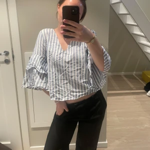 Randig blus med volangärm - Säljer en luftig blus med blå och vita vertikala ränder. Blusen har v-ringning, vida trekvartsärmar med volangdetalj och croppad passform. Perfekt för dig som gillar en avslappnad men trendig stil.