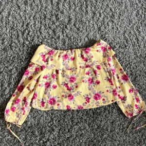 Gul blommig offshoulder blus NA-KD - Superfin gul blommig offshoulder blus från NA-KD x Andrea Hedenstedt. Blusen har volang upptill, långa ärmar med resår och knyt vid ärmslut. Perfekt för dig som vill sticka ut med färg och mönster. Säljes i storlek 34.