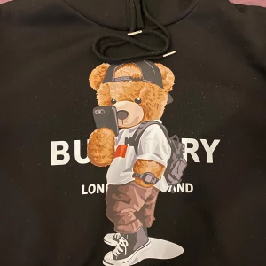 Svart hoodie med nalletryck från Burberry - Svart hoodie från Burberry med en stor, färgglad nalle som håller en mobil på bröstet. Tröjan har huva med snörning och klassisk känguruficka. Perfekt statement-plagg för dig som gillar streetstyle och coola prints.