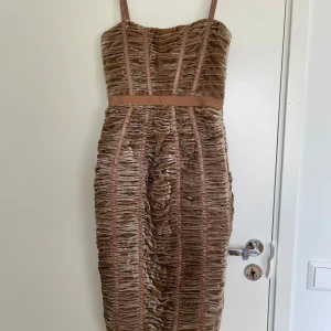 Beige sammetsklänning - Unik beige sammetsklänning med smala axelband och markerad midja. Klänningen har ett rynkat tyg med vertikala banddetaljer och dragkedja bak. Den är figurnära och har en lyxig känsla tack vare det glansiga sammetstyget. Perfekt för dig som vill sticka ut.