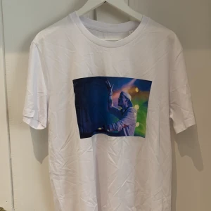 Vit Tim Bergling Foundation t-shirt - Vit t-shirt från Tim Bergling Foundation med tryck av Avicii på framsidan i färgstarka toner. Baksidan har en svart signatur och logga. Klassisk rund hals och korta ärmar, tillverkad i mjuk bomull. Perfekt för dig som gillar musik och vill ha en unik tee.