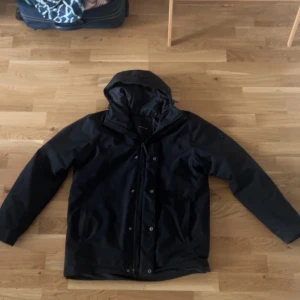 Svart vinterjacka Peak Performance - Svart vinterjacka från Peak Performance med HIPE-teknologi som gör den både vind- och vattentät. Jackan har huva med snörning, dragkedja och tryckknappar framtill samt praktiska fickor. Perfekt för kalla dagar och outdoor-äventyr.