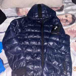 Mörkblå Peak Performance dunjacka - Säljer en mörkblå dunjacka från Peak Performance med huva och snyggt broderad logga på ärmen. Jackan har quiltad design, dragkedja framtill och ribbade muddar vid ärmslut. Perfekt för kalla dagar och har en cool, sportig vibe. Jackan är använd under en vinter och har en liten skada som ni kan se på bilden men ingeting man ser när den används. Perfekt nu till vintern som håller dig varm. Priset kan alltid diskutera så våga ge prisförslag.