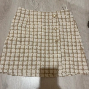 Beige och vit rutig kortkjol med pärlknappar - Superfin kortkjol i beige och vitt rutigt tyg med tre dekorativa pärlknappar framtill. Kjolen har en lätt A-linjeform och är gjord i ett strukturerat material som känns lyxigt. Perfekt för dig som gillar en chic och trendig stil.