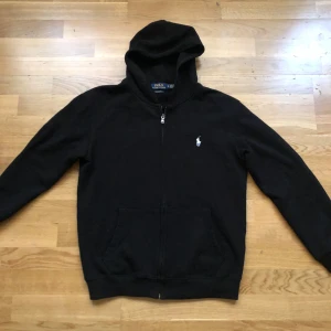 Svart hoodie från Polo Ralph Lauren - Svart hoodie från Polo Ralph Lauren med dragkedja framtill och klassisk vit logga på bröstet. Tröjan har huva, känguruficka och är långärmad. Custom fit-modell som passar perfekt till en avslappnad stil.