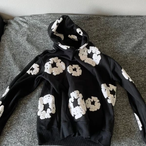 Svart hoodie från Denim Tears med tryck - Svart hoodie från Denim Tears med unika vita tryck i form av blommor över hela tröjan. Tröjan har huva och långa ärmar, tillverkad i mjuk bomull. Perfekt för dig som gillar streetwear och vill sticka ut med en cool design.
