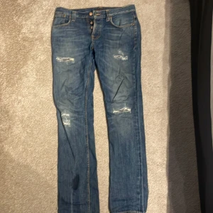 Blå jeans från Nudie med slitningar - Säljer ett par blå jeans från Nudie med snygga slitningar på framsidan och klassiska bakfickor med Nudie-logga. Jeansen har raka ben och knäppning framtill. Perfekta för dig som gillar en avslappnad och cool stil. Storlek 31/34